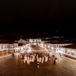 Praça histórica de Ouro Preto, iluminada à noite, com edifícios coloniais e pessoas circulando.