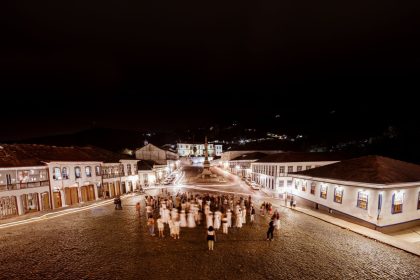 Praça histórica de Ouro Preto, iluminada à noite, com edifícios coloniais e pessoas circulando.