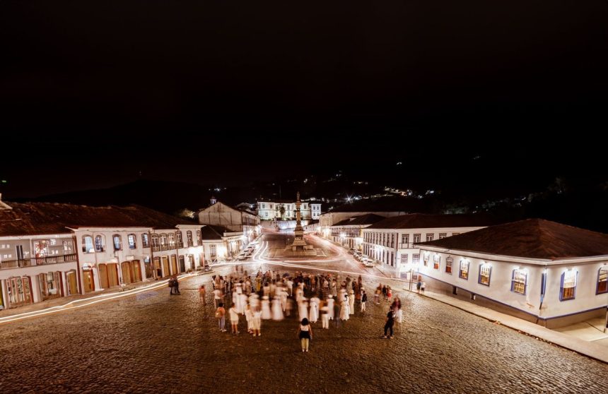 Praça histórica de Ouro Preto, iluminada à noite, com edifícios coloniais e pessoas circulando.
