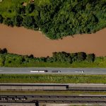 Vista aérea de caminhões trafegando por uma estrada às margens de um rio em Coronel Fabriciano, Minas Gerais