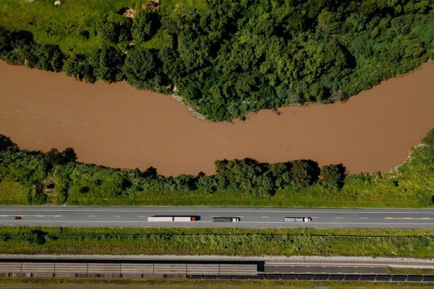 Vista aérea de caminhões trafegando por uma estrada às margens de um rio em Coronel Fabriciano, Minas Gerais