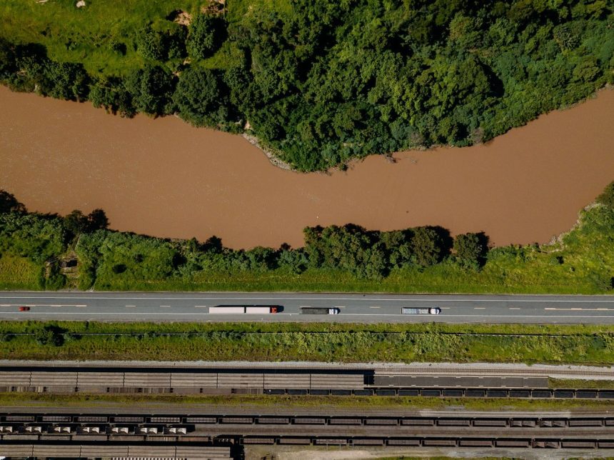 Vista aérea de caminhões trafegando por uma estrada às margens de um rio em Coronel Fabriciano, Minas Gerais