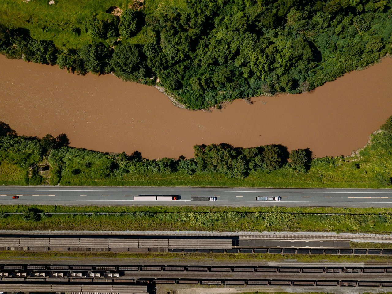 Vista aérea de caminhões trafegando por uma estrada às margens de um rio em Coronel Fabriciano, Minas Gerais